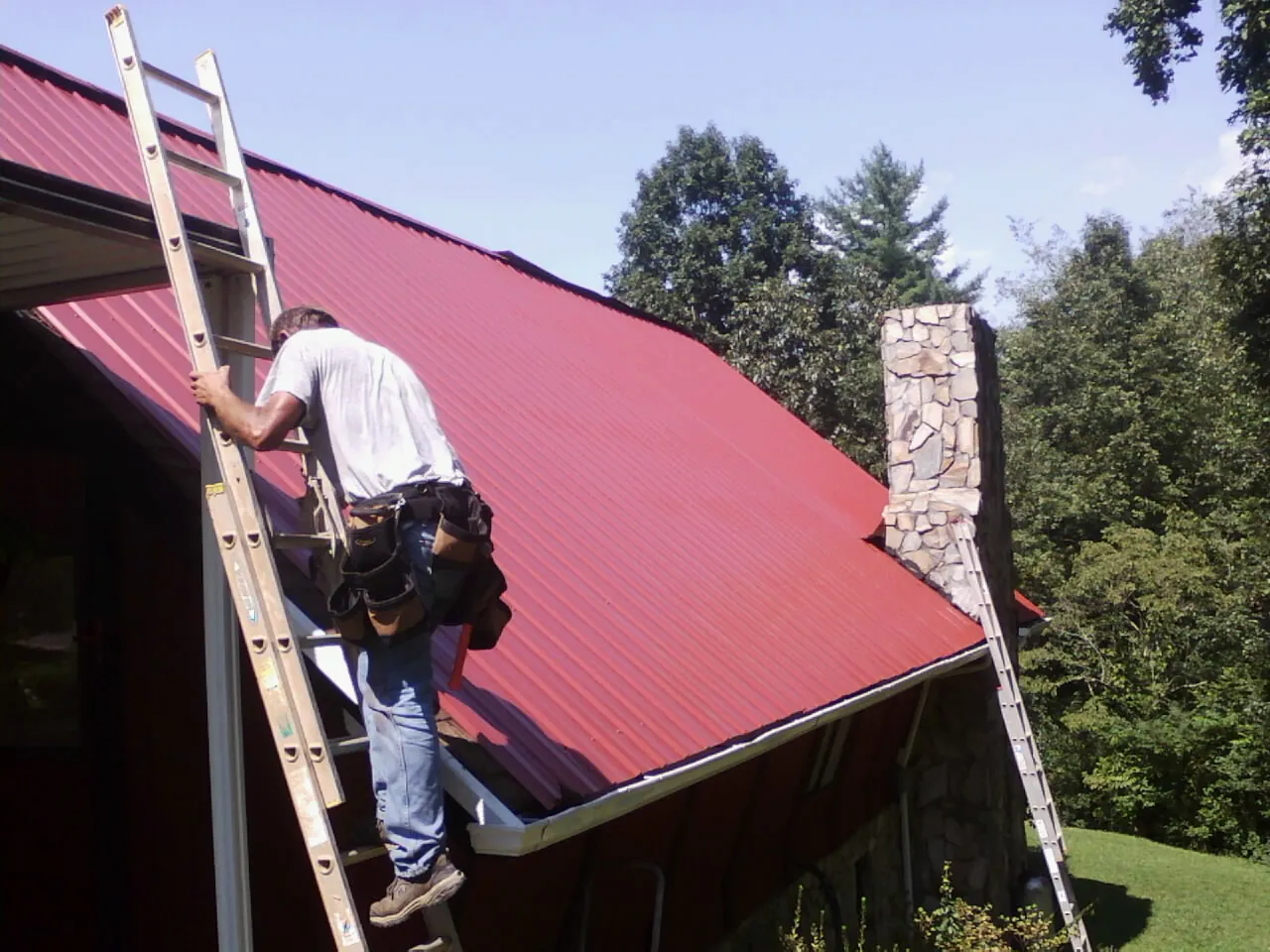 Roofer on a ladder beside a red metal roof and stone chimney — Soffit & Fascia Repair by Roofing Brothers in Wyckoff